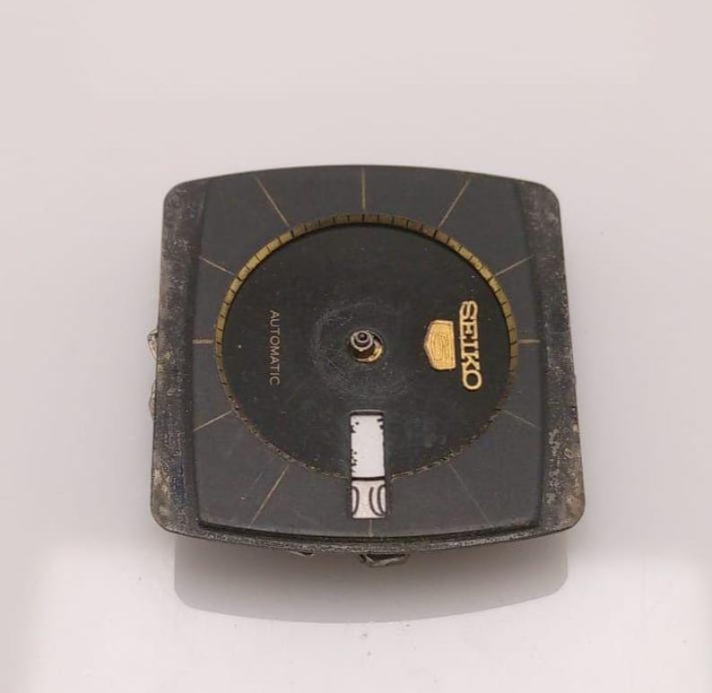 Puede incluir: Una esfera de reloj Seiko vintage con esfera negra y detalles dorados. La esfera cuadrada tiene una esfera circular en el centro. La palabra "AUTOMATIC" est&aacute; impresa en la esfera. El logotipo de Seiko es dorado. Es una pieza de repuesto.