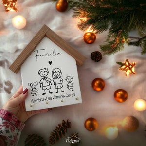 Op de afbeelding: Witte houten sleutelhanger in de vorm van een huis met het woord "Família" en een hart boven een gezin van vier stokfiguren. De namen Valentina, Luis, Simone en Giovanni staan onder de figuren.