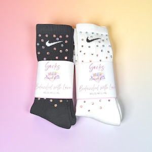 May include: Two pairs of socks, one black and one white, each adorned with sparkling embellishments. The socks feature the Nike swoosh logo and are wrapped with a label that reads "Socks" and "Bedazzled with Love."