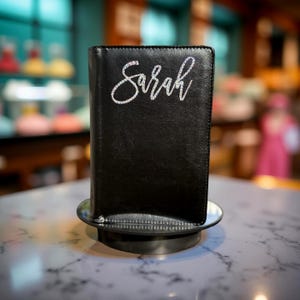 May include: Black leather journal with the name "Sarah" spelled out in sparkling rhinestones. The journal is displayed on a small, round metal stand, set against a blurred background. The journal appears to be new.