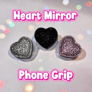 Könnte beinhalten: Drei herzförmige Handyhalterungen mit Spiegelfinish. Einer ist silber, einer schwarz und einer rosa. Der Text "Heart Mirror" und "Phone Grip" wird in rosa angezeigt.