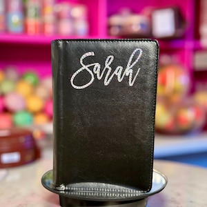 May include: A black leather journal with the name "Sarah" in silver script. The journal is closed and appears to be sitting on a small, round metal stand. The background is blurred, with hints of pink and colorful objects.