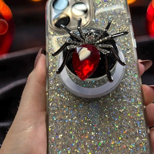 May include: A clear smartphone case with a silver glitter finish and the text CASE MATE printed at the bottom. Attached to the back is a circular phone grip featuring a black spider with a large, faceted red teardrop-shaped crystal body.
