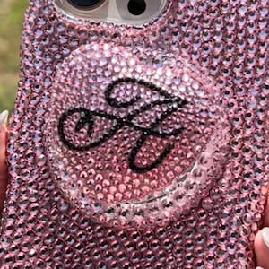 May include: Pink bedazzled phone case with a matching pop-up grip. The case is covered in small, sparkling rhinestones. The pop-up grip has a black cursive letter "A" on a clear background.