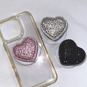 May include: Three heart-shaped phone grips in pink, silver, and black, each covered in sparkling rhinestones. The grips are on white bases, with a clear phone case in the background. A stylish accessory for any phone.