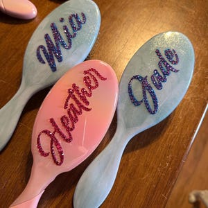 May include: Three personalized hairbrushes with names Mia, Heather, and Jade. The brushes have oval heads and long handles. Mia and Jade are on light blue brushes, and Heather is on a pink brush. The names are written in sparkly, decorative lettering.