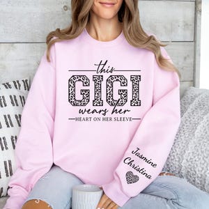 May include: Pink sweatshirt with the text "This Gigi wears her heart on her sleeve" in black lettering with a leopard print design. The text "Jasmine Christina" is on the sleeve with a small heart.