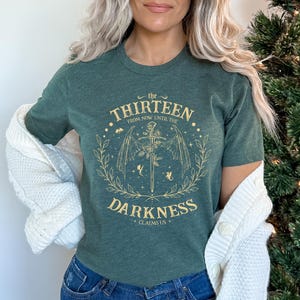 May include: A dark green t-shirt with a gold graphic design featuring a sword and wings. The text on the shirt reads "The Thirteen From Now Until The Darkness Claims Us."