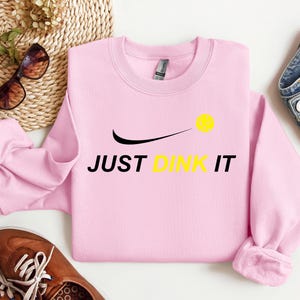 Może przedstawiać: R&oacute;żowa bluza z czarnym swoosh i napisem "JUST DINK IT" w kolorze czarnym. Ż&oacute;łta piłka do pickleballa znajduje się nad swoosh.