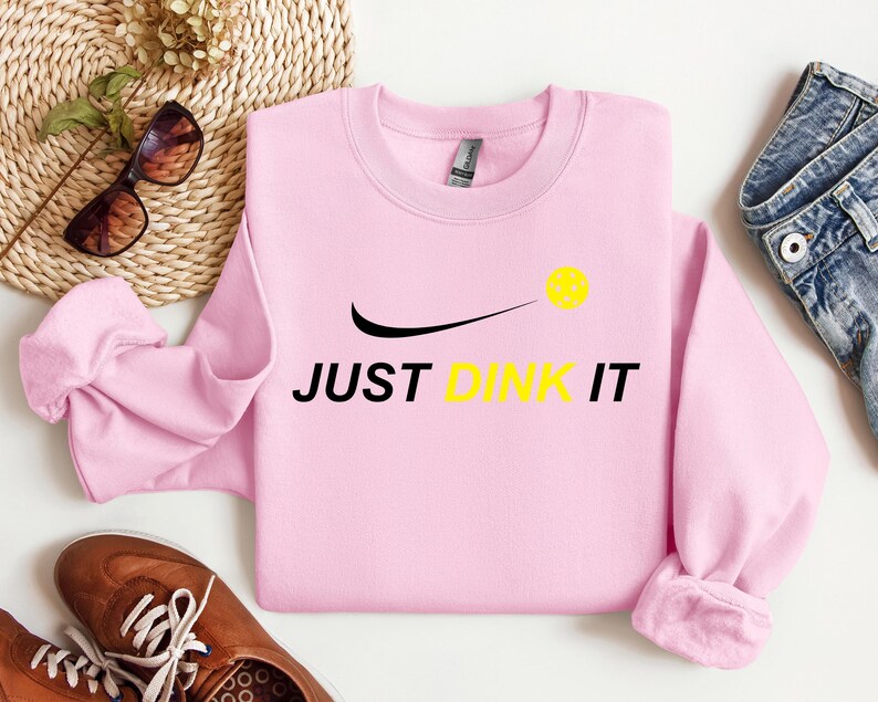 Może przedstawiać: R&oacute;żowa bluza z czarnym swoosh i napisem "JUST DINK IT" w kolorze czarnym. Ż&oacute;łta piłka do pickleballa znajduje się nad swoosh.