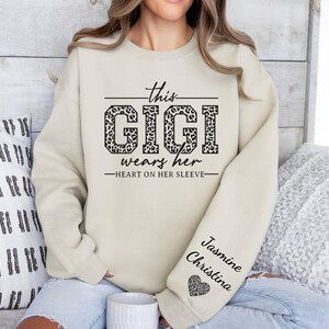 May include: A beige sweatshirt with the text "This Gigi wears her heart on her sleeve" in black. The word "Gigi" is printed in a leopard print pattern. The sleeve has the names "Jasmine Christina" and a heart with a leopard print pattern.