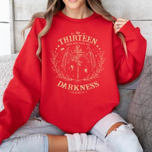 May include: Red sweatshirt with a gold graphic design featuring a sword, wings, and the text "the thirteen from now until the darkness claims us".