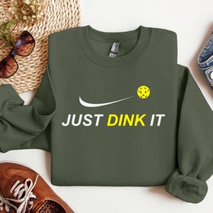Może przedstawiać: Zielony sweter z białym swoosh i napisem "JUST DINK IT" w kolorze białym. Ż&oacute;łta piłka do pickleballa znajduje się nad swoosh.