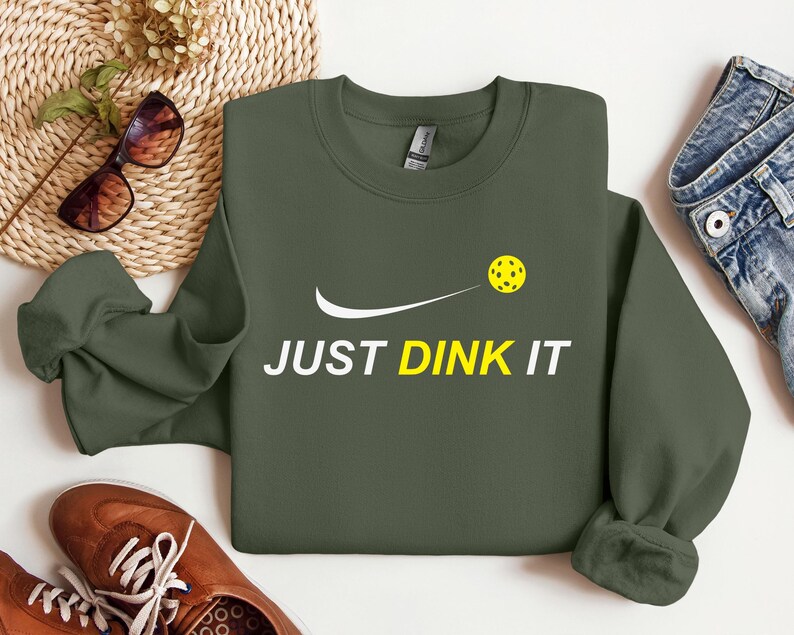 Może przedstawiać: Zielony sweter z białym swoosh i napisem "JUST DINK IT" w kolorze białym. Ż&oacute;łta piłka do pickleballa znajduje się nad swoosh.
