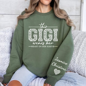 May include: Green sweatshirt with the text "This Gigi wears her heart on her sleeve" in white with a leopard print design. The sleeve has the names "Jasmine Christina" in white with a heart with a leopard print design.