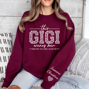 May include: Burgundy sweatshirt with white text that reads "This Gigi wears her heart on her sleeve." The text is surrounded by a leopard print design. The sleeve has the names "Jasmine" and "Christina" written in white with a pink heart.