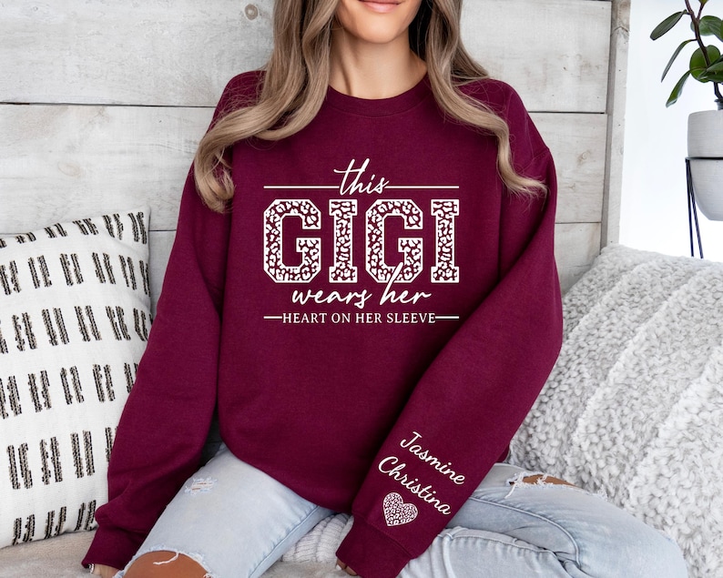 May include: Burgundy sweatshirt with white text that reads "This Gigi wears her heart on her sleeve." The text is surrounded by a leopard print design. The sleeve has the names "Jasmine" and "Christina" written in white with a pink heart.