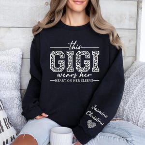 May include: Black sweatshirt with white text that reads "This Gigi wears her heart on her sleeve". The text is in a leopard print font. The sleeve has the name "Jasmine Christina" written in white text with a small heart.
