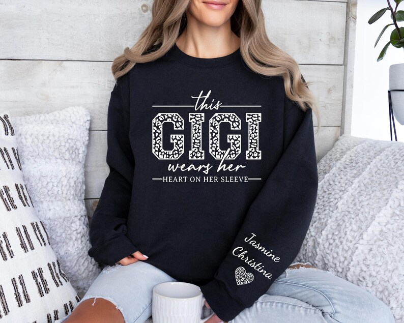 May include: Black sweatshirt with white text that reads "This Gigi wears her heart on her sleeve". The text is in a leopard print font. The sleeve has the name "Jasmine Christina" written in white text with a small heart.