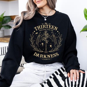 May include: Black sweatshirt with a gold graphic print of a winged sword and floral design. The text reads "The Thirteen From Now Until The Darkness Claims Us."