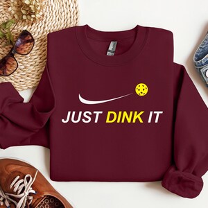 Może przedstawiać: Bordowy sweter z białym swoosh i napisem "JUST DINK IT" w kolorze białym. Ż&oacute;łta piłka do pickleballa leci przez swoosh.
