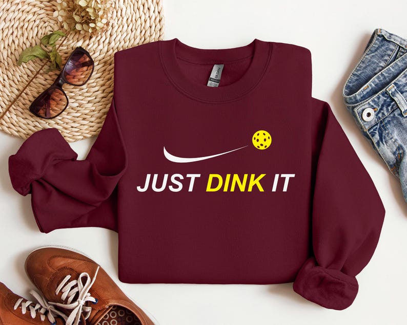 Może przedstawiać: Bordowy sweter z białym swoosh i napisem "JUST DINK IT" w kolorze białym. Ż&oacute;łta piłka do pickleballa leci przez swoosh.
