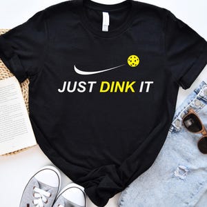 Może przedstawiać: Czarny T-shirt z białym swoosh i ż&oacute;łtą piłką do pickleballa. Tekst "JUST DINK IT" jest nadrukowany na biało pod piłką do pickleballa.