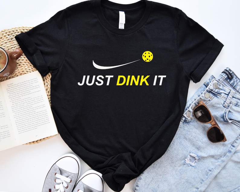 Może przedstawiać: Czarny T-shirt z białym swoosh i ż&oacute;łtą piłką do pickleballa. Tekst "JUST DINK IT" jest nadrukowany na biało pod piłką do pickleballa.