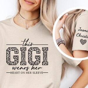 May include: Beige t-shirt with a leopard print graphic that says "This Gigi wears her heart on her sleeve". The sleeve of the shirt has a leopard print heart with the names "Jasmine Christina" written above it.