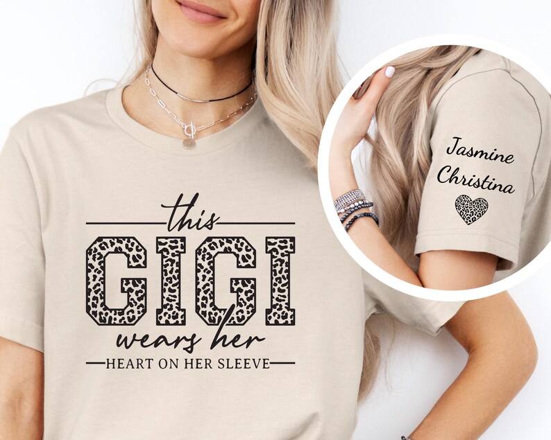 May include: Beige t-shirt with a leopard print graphic that says "This Gigi wears her heart on her sleeve". The sleeve of the shirt has a leopard print heart with the names "Jasmine Christina" written above it.