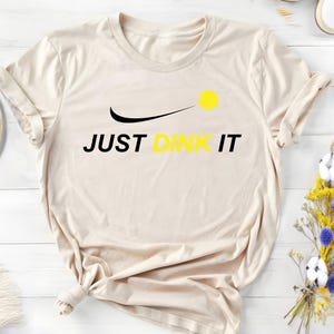 Może przedstawiać: Beżowa koszulka z czarnym swoosh i napisem "JUST DINK IT" w kolorze czarnym. Ż&oacute;łta piłka do pickleballa znajduje się nad swoosh.