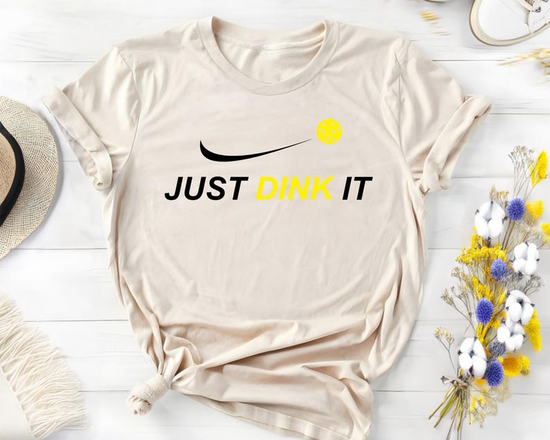 Może przedstawiać: Beżowa koszulka z czarnym swoosh i napisem "JUST DINK IT" w kolorze czarnym. Ż&oacute;łta piłka do pickleballa znajduje się nad swoosh.