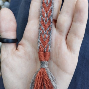 Könnte beinhalten: Nahaufnahme einer Hand, die eine gewebte Schnur mit einem roten und grauen Muster hält. Die Schnur hat an einem Ende einen Quasten.