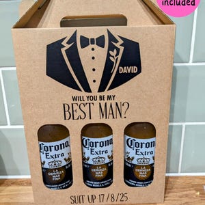 May include: A brown cardboard box with a tuxedo design and the text "WILL YOU BE MY BEST MAN?" and "SUIT UP 17/8/25". The box has three slots for bottles of Corona Extra beer. The text "Beer not included" is written on a pink circle in the upper right corner of the image.