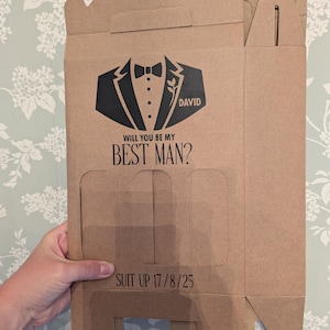 May include: A brown cardboard box with a tuxedo graphic and the text "Will you be my Best Man?" and "Suit Up 17/8/25". The box has a flap on the top and a cutout on the front.