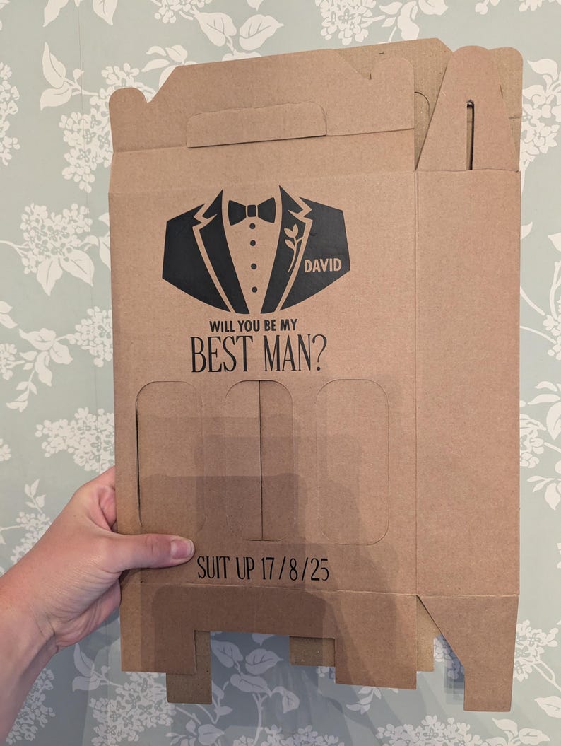 May include: A brown cardboard box with a tuxedo graphic and the text "Will you be my Best Man?" and "Suit Up 17/8/25". The box has a flap on the top and a cutout on the front.