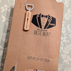 May include: A brown cardboard box with a black tuxedo graphic and the text "Will you be my Best Man?" and "Suit Up 29/05/2026". A wooden bottle opener with the text "Daniel Best Man" is attached to the box.