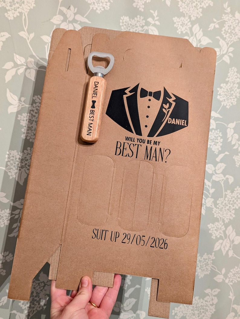 May include: A brown cardboard box with a black tuxedo graphic and the text "Will you be my Best Man?" and "Suit Up 29/05/2026". A wooden bottle opener with the text "Daniel Best Man" is attached to the box.