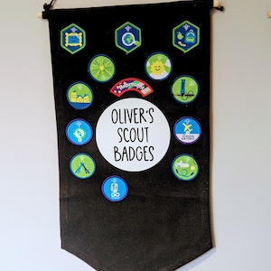 May include: A black banner displaying a collection of colourful scout badges. The banner is suspended from a wooden dowel with a black cord. The text "Oliver's Scout Badges" is centrally positioned. The badges feature diverse shapes and designs.