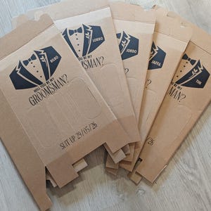 May include: Five brown cardboard boxes with a tuxedo graphic and the text "Will you be my groomsman?" and "Suit up 29/05/26".