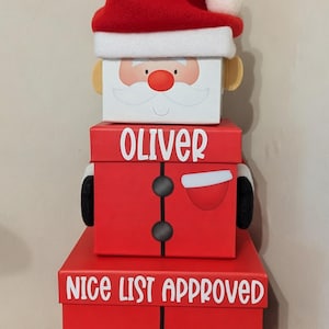 May include: A festive stack of three boxes designed as Santa Claus. The top box features a Santa face and hat. The middle box is labelled "OLIVER". The bottom box reads "NICE LIST APPROVED".