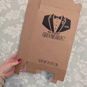 May include: A brown cardboard box with a black tuxedo graphic and the text "Will you be my groomsman?" and "Suit up 29/05/26". The box also has the name "Bilbo" printed on it.