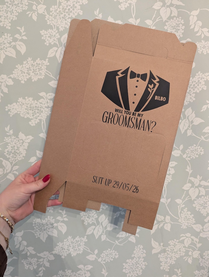 May include: A brown cardboard box with a black tuxedo graphic and the text "Will you be my groomsman?" and "Suit up 29/05/26". The box also has the name "Bilbo" printed on it.