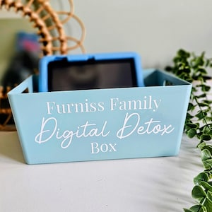 May include: A pale blue storage box with the text "Furniss Family Digital Detox Box" in white script. A blue tablet is inside the box. The box is on a white surface, with greenery visible in the background.