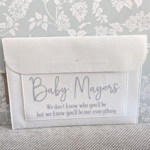 May include: A white felt baby keepsake holder with a flap closure. The front has a rectangular pocket with the text "Baby Mayers" and the phrase "We don't know who you'll be but we know you'll be our everything" in grey script.