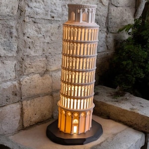 Puede incluir: Un modelo de cerámica blanca de la Torre Inclinada de Pisa con una luz en el interior, iluminando el interior. El modelo está sobre una base negra y está sentado sobre una superficie de piedra.