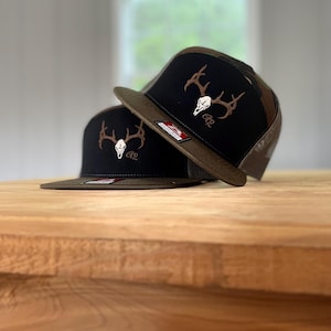 Könnte beinhalten: Zwei schwarze Trucker-Caps mit braunem Schirm und einem Camouflage-Muster auf der Oberseite. Jede Cap hat ein gesticktes Hirschschädel- und Geweih-Design in Braun und Weiß.