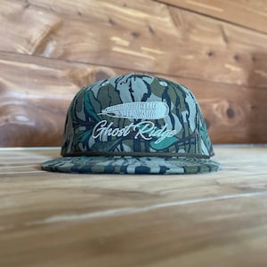 May include: A camouflage baseball cap with a flat brim. The cap features a white embroidered feather design above the words "Ghost Ridge" in a script font. The hat is primarily green, brown, and tan.