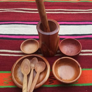 Puede incluir: Un juego de utensilios de cocina de madera, que incluye un mortero y un pilón, tres cucharas y cuatro cuencos. Los utensilios son todos de madera marrón claro y están dispuestos sobre una tela de rayas rojas y blancas.