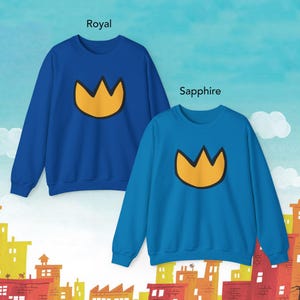 May include: Two crewneck sweatshirts, one royal blue and one sapphire blue, each featuring a gold crown graphic. The word "Royal" is above the blue sweatshirt, and "Sapphire" is above the other. The background has a cityscape.